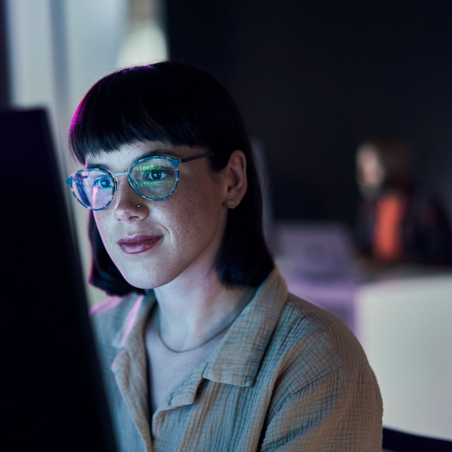A woman with short dark hair and glasses is focused on a computer screen in a dimly lit office. A blurred colleague works in the background.