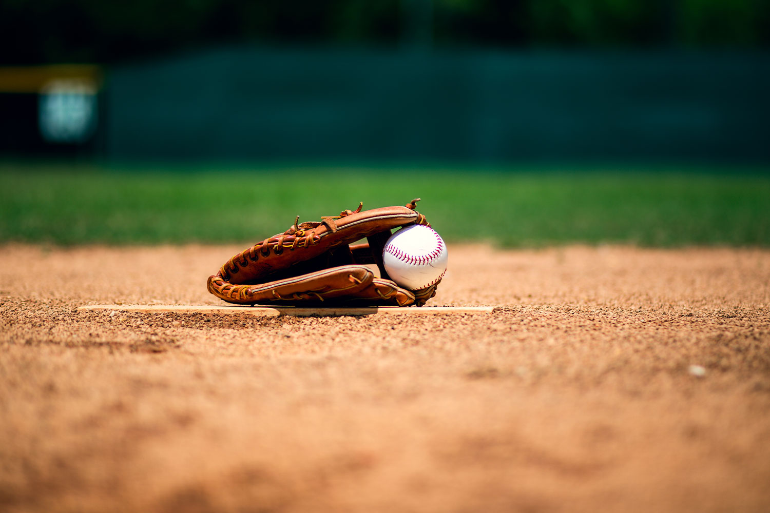 A baseball glove with a baseball inside it, lying on a pitcher's mound on a baseball field, with the green outfield blurred in the background.