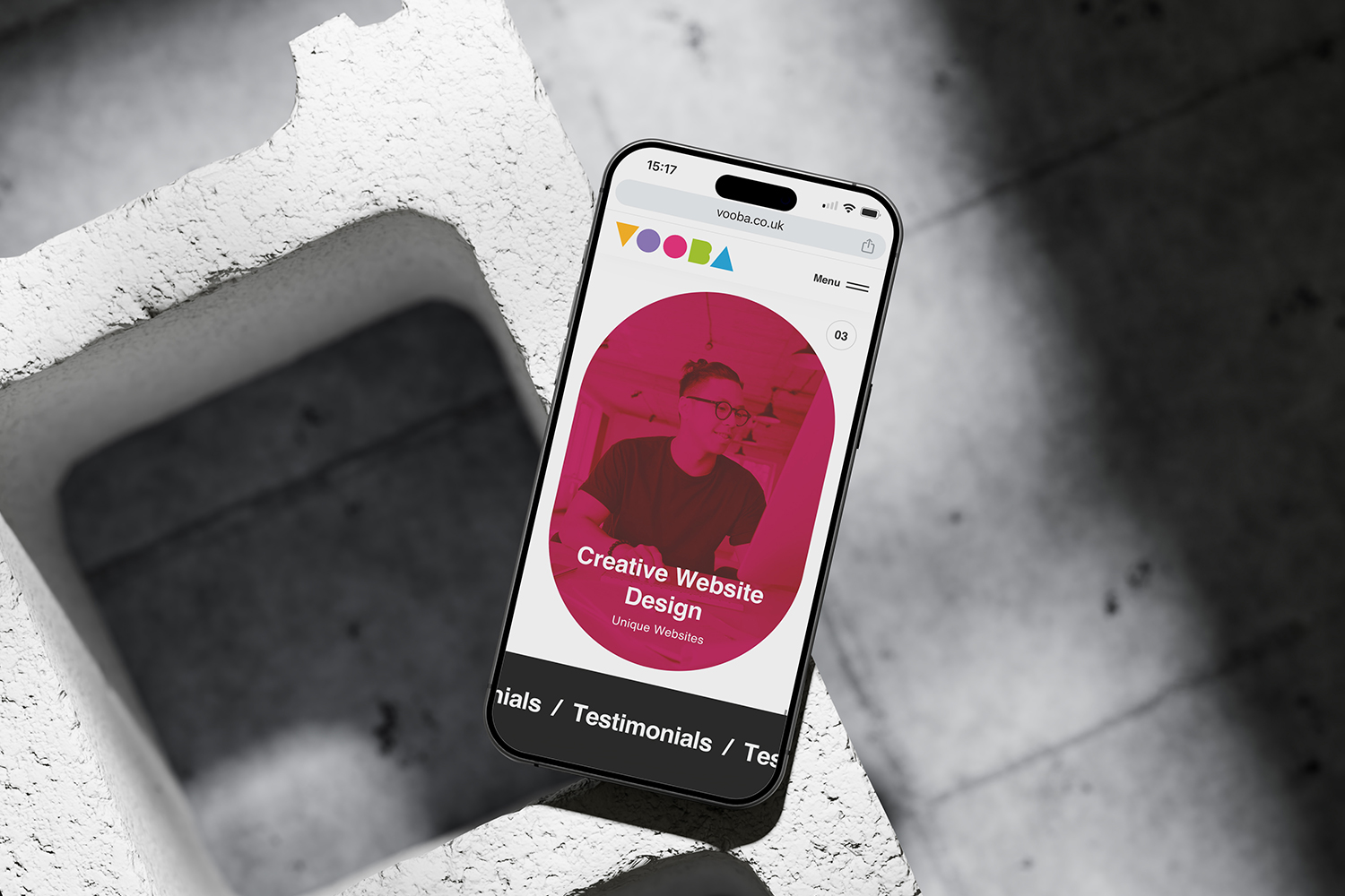 A smartphone showing a colorful website design with a creative layout. The phone is placed on a concrete block, casting a shadow on the surface. The image is likely used as a header or main visual for a web design showcase.t