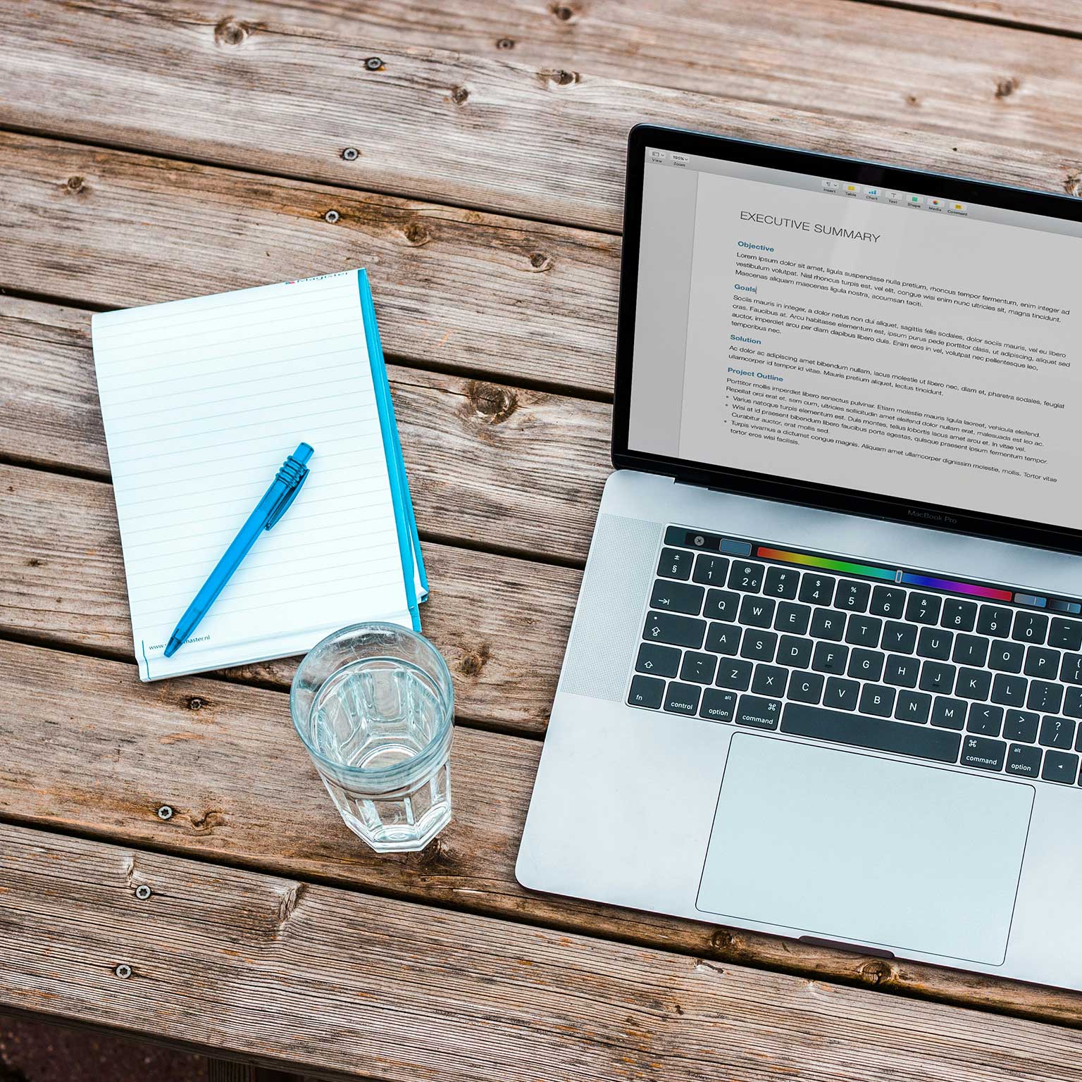 Top view of a workspace on a wooden table, featuring an open laptop with an 'Executive Summary' on the screen, a notepad with a blue pen on it, and a glass of water, suggesting a productive remote work setting.