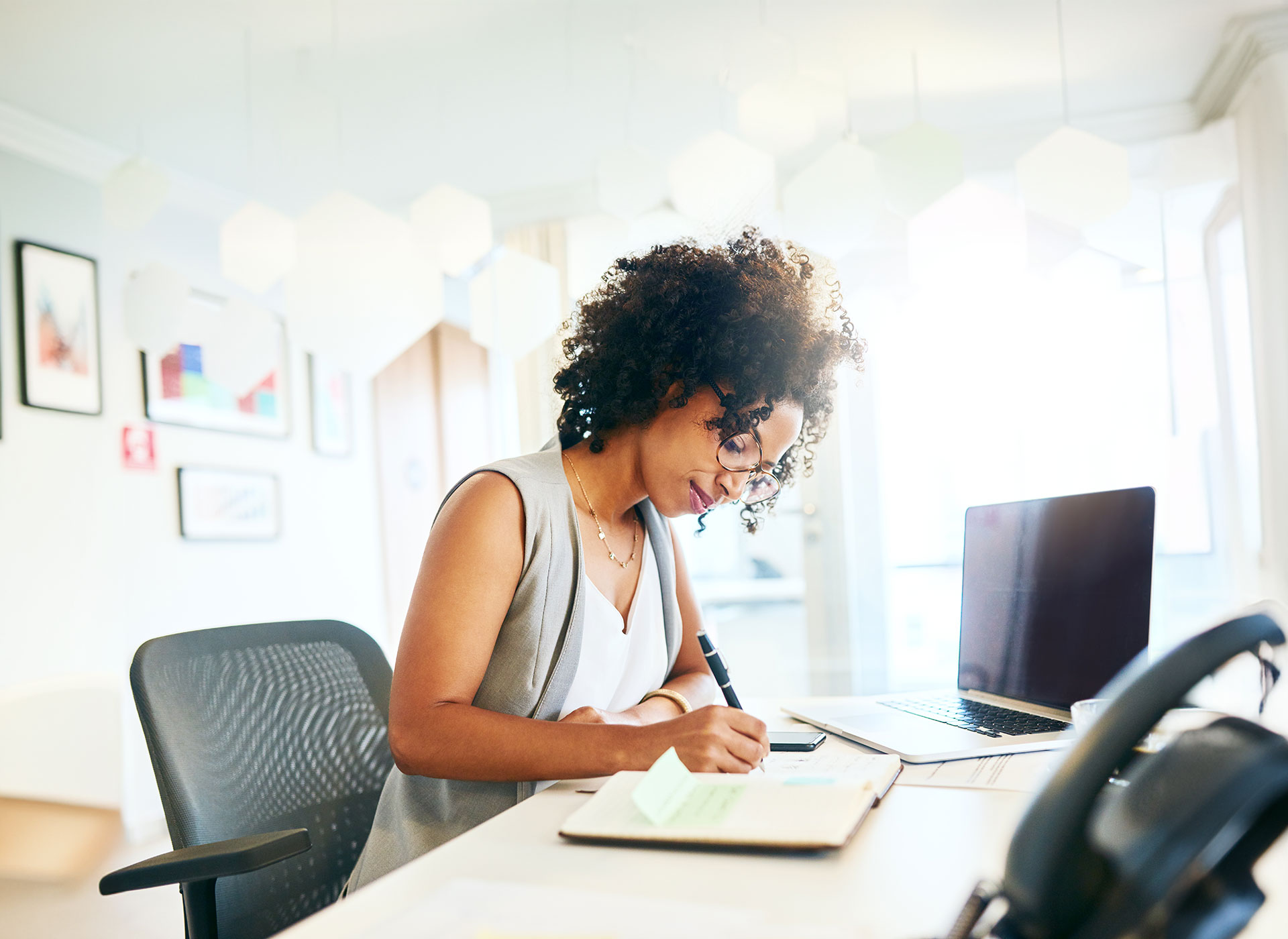 Image of a focused African American woman working at a modern office desk, writing notes while looking at her laptop. The office is brightly lit, with decorative geometric shapes hanging from the ceiling and artwork on the walls.