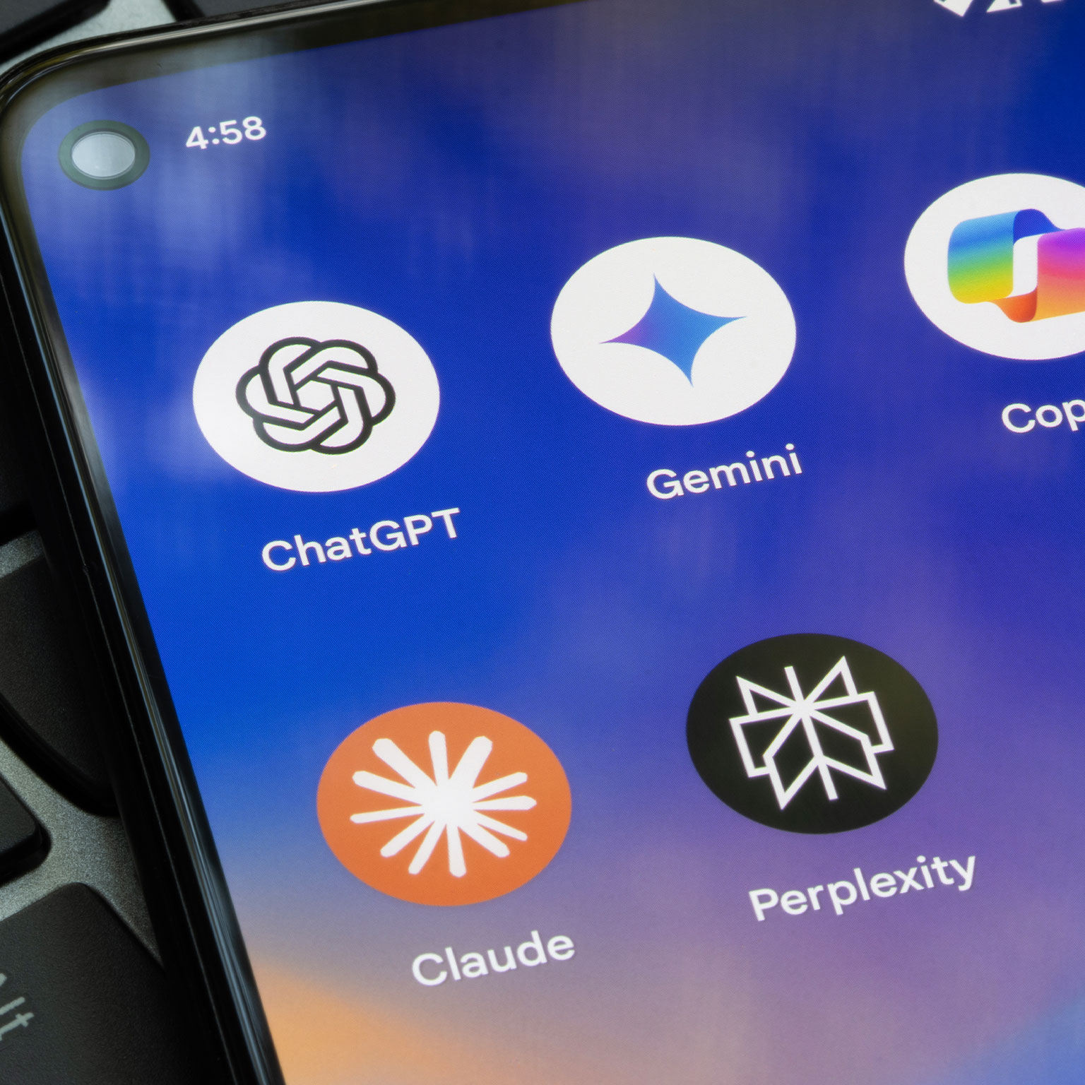 Close-up of a smartphone screen displaying AI app icons: ChatGPT, Gemini, Copilot, Claude, and Perplexity. Phone rests on a laptop keyboard.