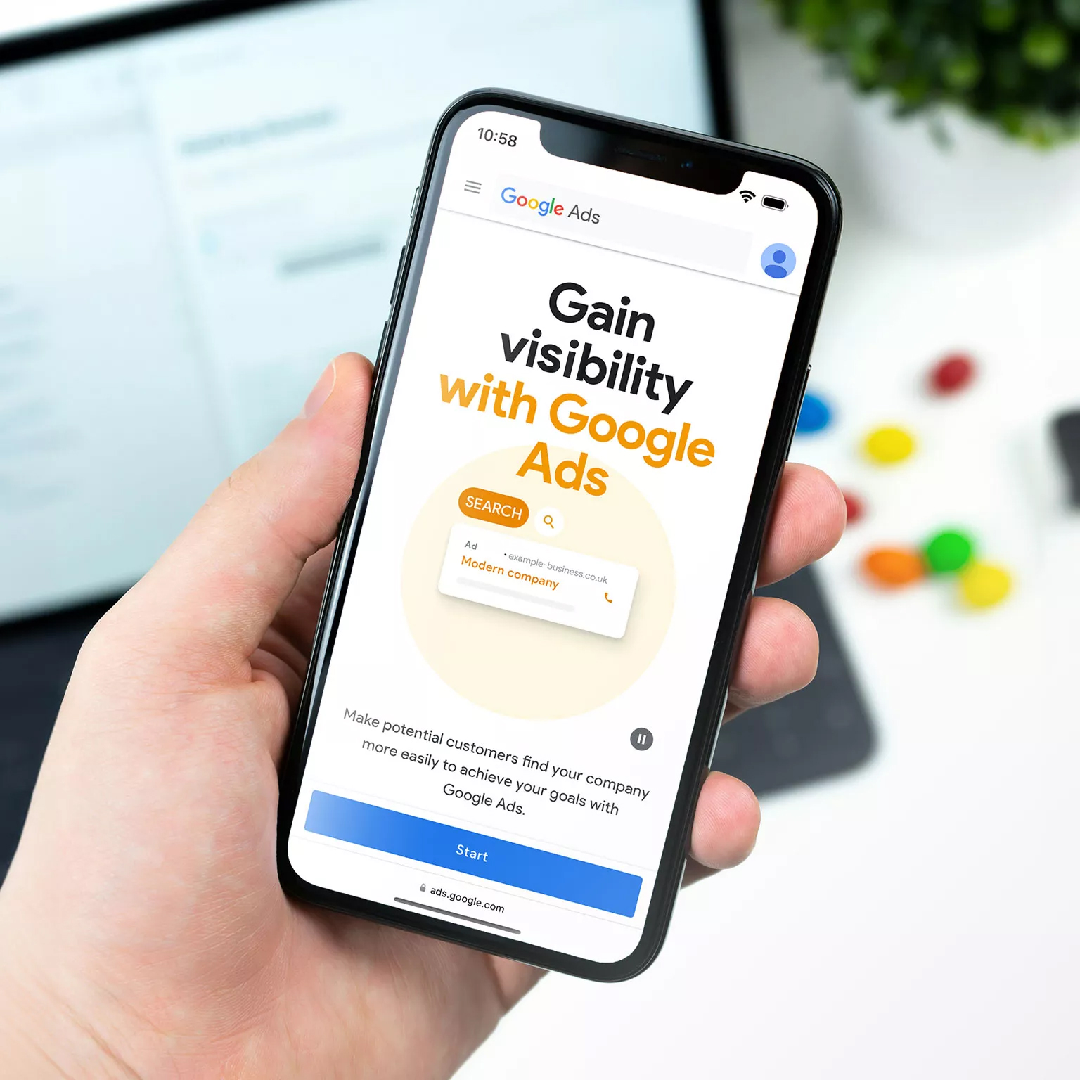 Close-up of a person's hand holding a smartphone displaying a Google Ads advertisement on the screen, promoting visibility for businesses through search advertising, with a mock search result for a 'modern company' and a 'Start' button at the bottom.