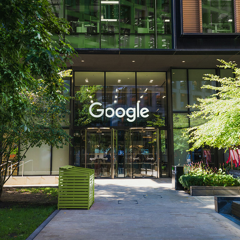 Google office entrance with a large logo on the glass facade, surrounded by greenery.