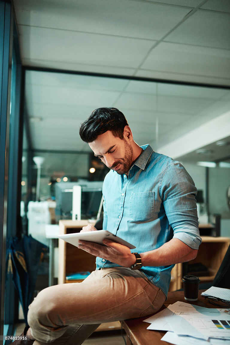 Man in a casual blue shirt using a tablet while sitting on a desk in an office setting.