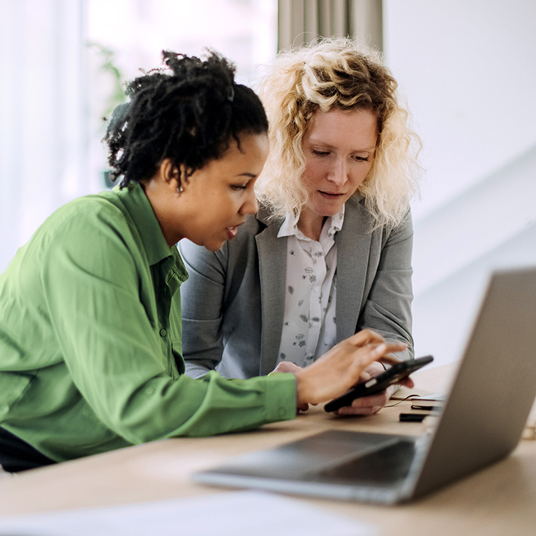Two businesswomen collaborating at a desk, one showing the other something on a smartphone, with a laptop in front of them.