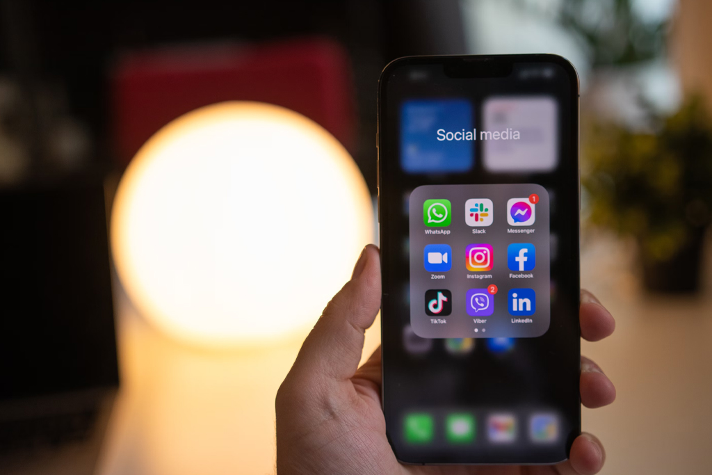 Hand holding a smartphone displaying social media apps including WhatsApp, Slack, Messenger, Zoom, Instagram, Facebook, TikTok, Viber, and LinkedIn.