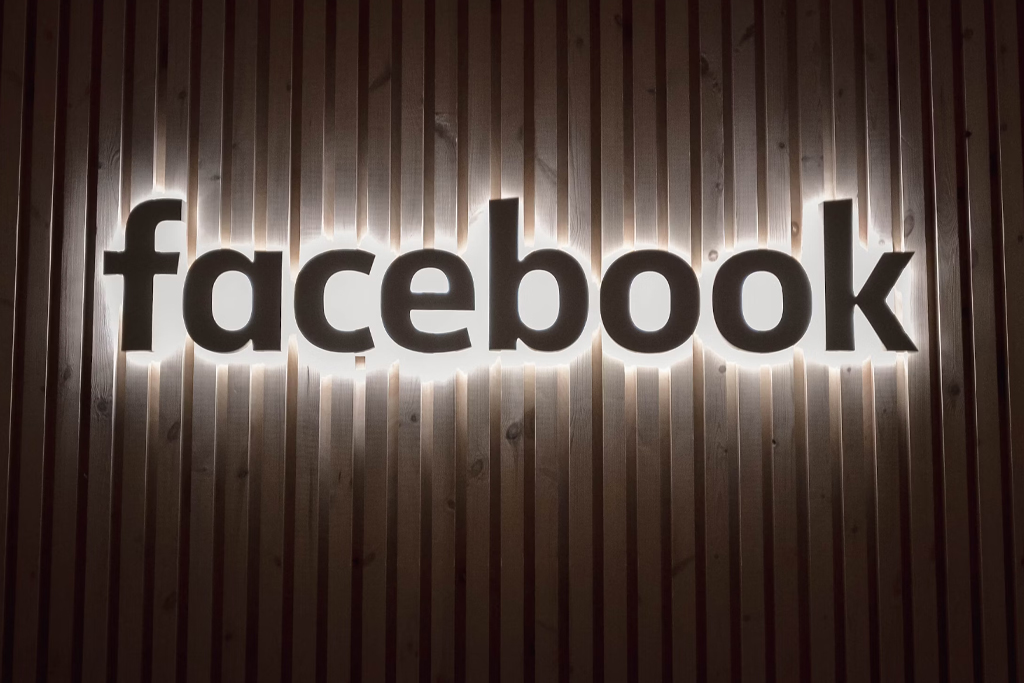 Neon sign of the Facebook logo mounted on a wooden slatted wall, emitting a warm glow in a dark setting.