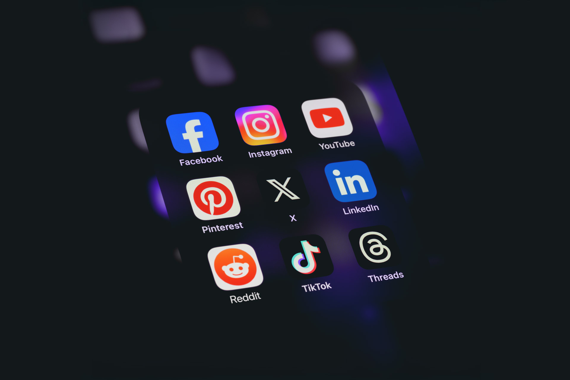 Smartphone screen displaying a collection of popular social media app icons including Facebook, Instagram, YouTube, Pinterest, LinkedIn, Reddit, TikTok, and Threads, glowing against a dark background.