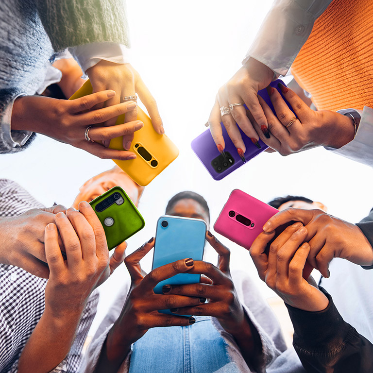 Group of diverse hands holding smartphones with colorful cases reaching towards the center, symbolizing social connectivity and digital interaction.