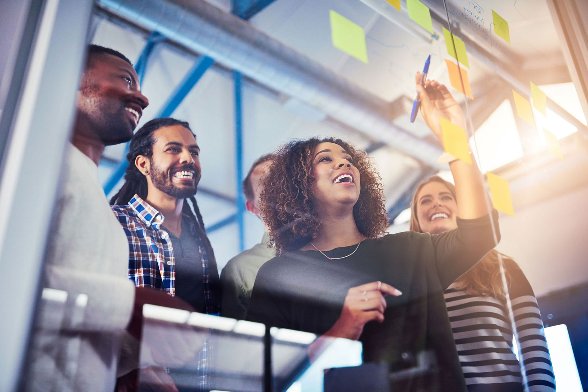 Diverse group of business professionals collaborating in a bright office environment, enthusiastically engaging in a brainstorming session with sticky notes on a glass wall.